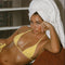 Woman in yellow bikini with a towel on her head, holding a drink, in a room with a window.