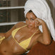 Woman in yellow bikini with a towel on her head, holding a drink, in a room with a window.